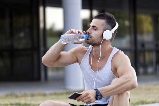 Портрет молодого человека, пьющего воду из бутылки во время сидения и отдыха после тренировки
. - Фото, изображение