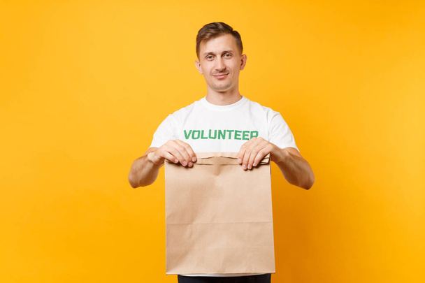 Portrait of man in white t-shirt written inscription green title volunteer hold blank craft paper bag for takeaway isolated on yellow background. Voluntary free assistance help charity grace concept - Valokuva, kuva