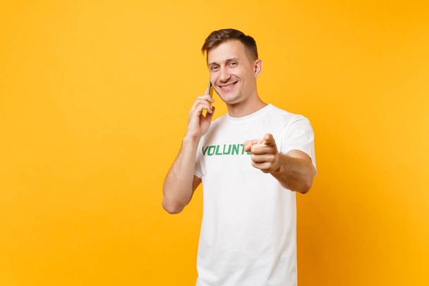 Retrato de homem jovem em camiseta branca com inscrição escrita título verde voluntário falando no celular isolado em fundo amarelo. Assistência gratuita voluntária ajuda, caridade conceito de trabalho de graça
 - Foto, Imagem