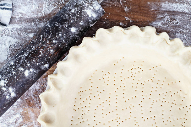 Homemade butter pie crust in pie plate with fluted pinched edge, rolling pin and extra ball of dough over floured rustic wooden background. Crust has been perforated with fork and ready for baking. - Photo, image