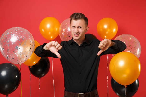 Dissatisfied young man in black classic shirt showing thumbs down on bright red background air balloons. St. Valentine's International Women's Day Happy New Year birthday mockup holiday party concept - Photo, Image
