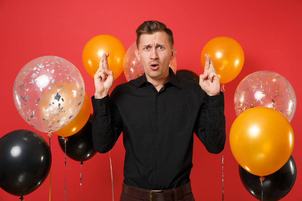 Παρακαλεί νεαρός άνδρας στο κλασικό πουκάμισο κρατώντας τα δάχτυλα διέσχισαν, περιμένει ιδιαίτερη στιγμή σε κόκκινο φόντο αερόστατα. Παγκόσμια ημέρα της γυναίκας, ευτυχισμένο το νέο έτος γενεθλίων mockup holiday κόμμα έννοια - Φωτογραφία, εικόνα