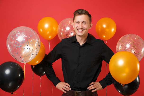 Smiling young man in black classic shirt standing with arms akimbo on bright red background air balloons. Valentine's, International Women's Day, Happy New Year, birthday mockup holiday party concept - Photo, Image