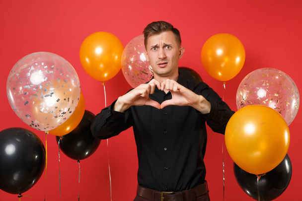 Puzzled young man in black classic shirt showing heart gesture on bright red background air balloons. St. Valentine's, International Women's Day, Happy New Year birthday mockup holiday party concept - Foto, Imagem