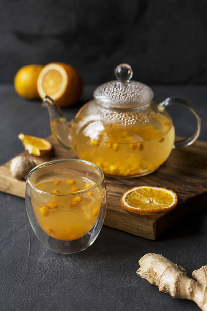 Hot sea buck thorn tea in glass teapot with glass cup, orange and ginger near, at wooden board on black background - Foto, Imagen