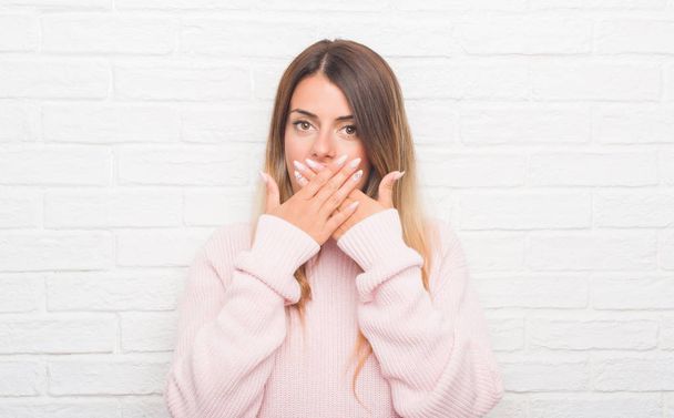Jonge volwassen vrouw over witte bakstenen muur dragen winter outfit thuis geschokt bedekking mond met handen voor fout. Geheime concept. - Foto, afbeelding