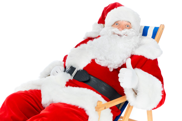 бородатий Санта Клаус показує великий палець вгору і сидить у пляжному кріслі ізольовано на білому
 - Фото, зображення