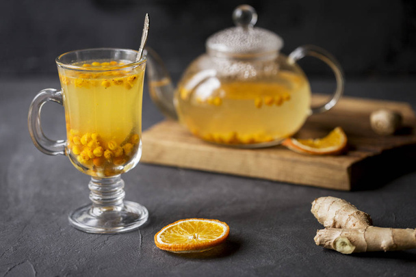 Hot sea buck thorn tea in glass teapot with glass cup and spoon, orange and ginger near, at wooden board on black background - Photo, Image
