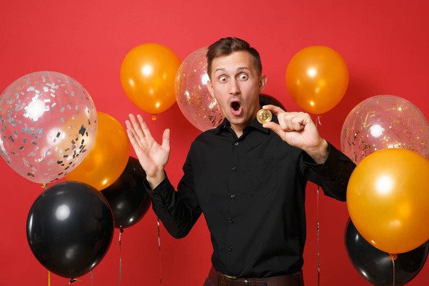 Shocked young man in classic shirt spreading hands, holding bitcoin, metal coin of golden color, future currency on red background air balloons. Happy New Year, birthday mockup holiday party concept - Photo, Image