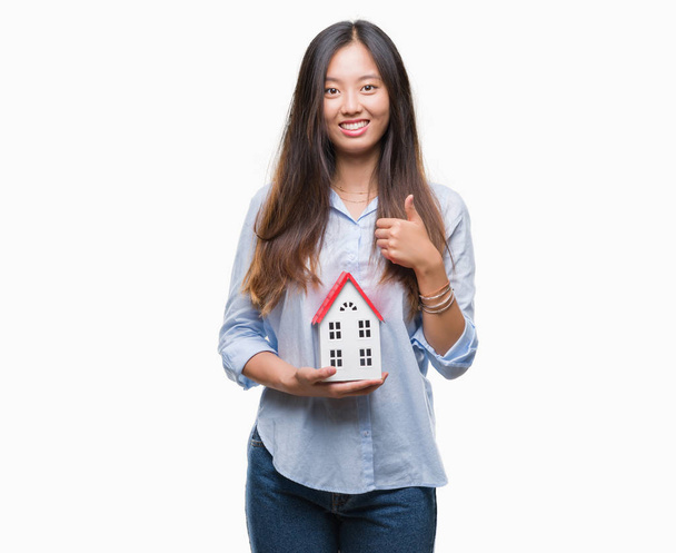 Jonge Aziatische vrouw echte staat agent houden huis geïsoleerd achtergrond blij met de grote glimlach doen ok teken, duim omhoog met vingers, uitstekende teken - Foto, afbeelding