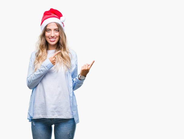 分離背景笑顔で側に 2 つの手と指で指しているカメラ目線の上クリスマス帽子身に着けている美しい若いブロンドの女性. - 写真・画像