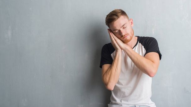 junger rothaariger Mann über grauer Grunge-Wand schläft müde träumend und posiert mit Händen zusammen, während er mit geschlossenen Augen lächelt. - Foto, Bild