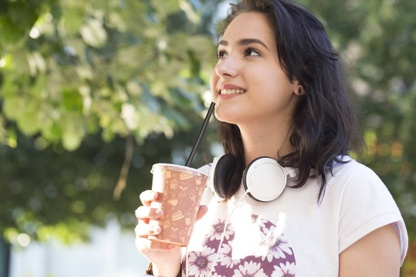 Portrait of a beautiful young teenage girl with headphones around her neck outdoors. She was holding a large plastic cup with juice. With a beautiful smile and joyful emotions. Copy space - Photo, Image