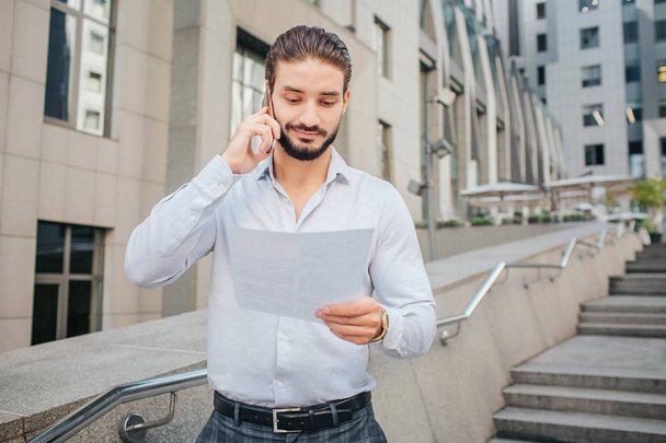 Handsome and positive young man stands on steps. He holds pece of paper and smiles. Businessman talks on phone. He looks at document. - Foto, imagen