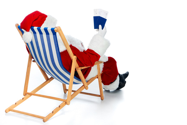 Rückansicht des Weihnachtsmannes mit zwei Pässen und Flugtickets für die Weihnachtsreise, während er im Strandkorb isoliert auf weiß sitzt  - Foto, Bild