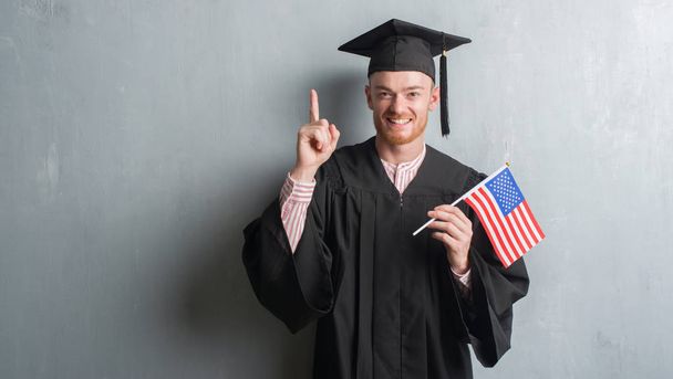Jonge roodharige man over grijs grunge muur dragen afgestudeerde uniform holding Amerika vlag verrast met een idee of vraag wijzende vinger met blij gezicht, nummer één - Foto, afbeelding