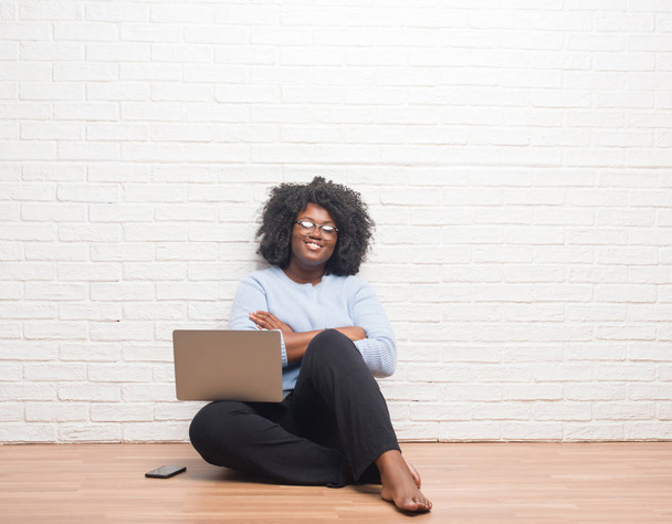 Jeune femme afro-américaine assise sur le sol à l'aide d'un ordinateur portable à la maison heureux visage souriant avec les bras croisés regardant la caméra. Personne positive
. - Photo, image