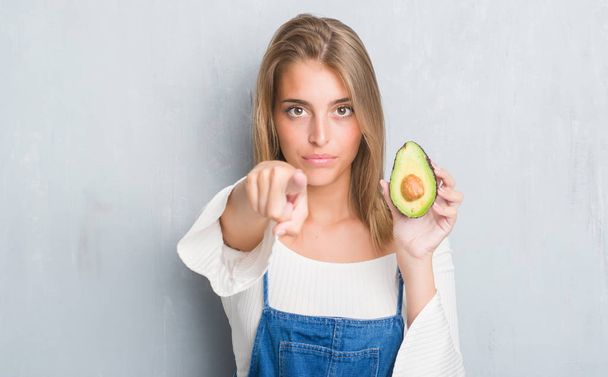 Mooie jonge vrouw over grunge grijs muur eten avocado wijzen met de vinger naar de camera en u, hand ondertekenen, positieve en zelfverzekerd gebaar van de voorkant - Foto, afbeelding
