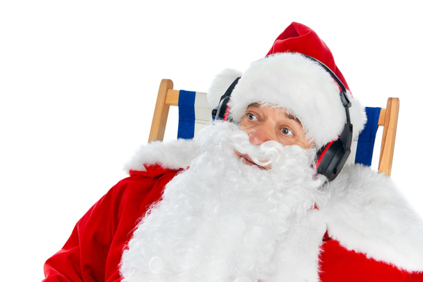 Weihnachtsmann hört Musik mit Kopfhörern, während er im Strandkorb isoliert auf weißem Grund sitzt - Foto, Bild