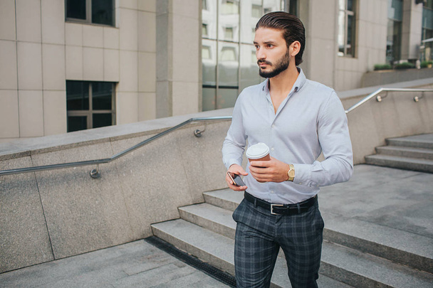 Bearded and handsome young man stands on steps and poses. He looks straight in front of hinself. Businessman holds phone and cup of hot drink. He looks serious. - Foto, immagini