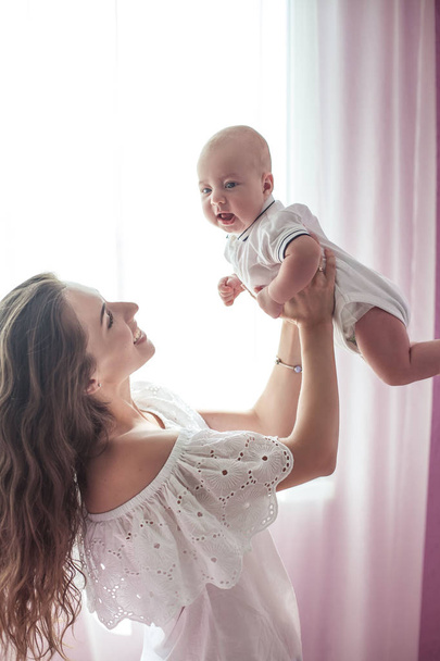 Kaunis nuori äiti pitää vauvaa sylissään. Äiti pitää vauvan jalkoja käsissään 3 kuukautta. Rakkaudesta. Äitiys. Onnellisuus. Talo
. - Valokuva, kuva