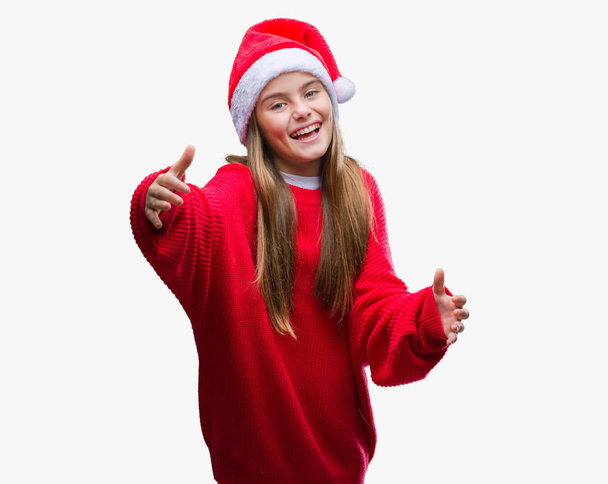Jeune belle fille portant un chapeau de Noël sur fond isolé regardant la caméra souriant à bras ouverts pour un câlin. Expression joyeuse embrassant le bonheur
. - Photo, image