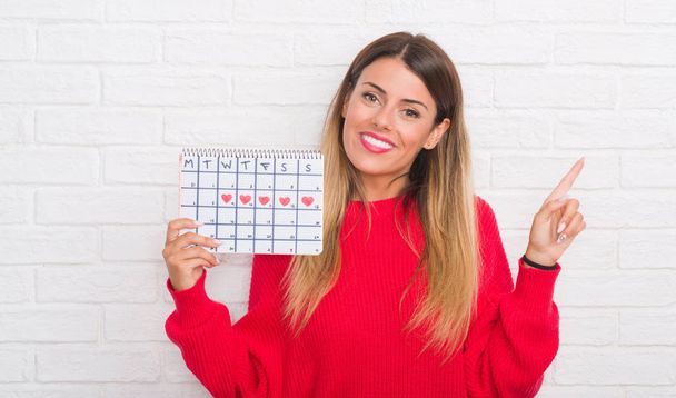 junge erwachsene Frau über weiße Backsteinmauer mit Zeitrahmen Kalender sehr glücklich zeigt mit Hand und Finger zur Seite - Foto, Bild