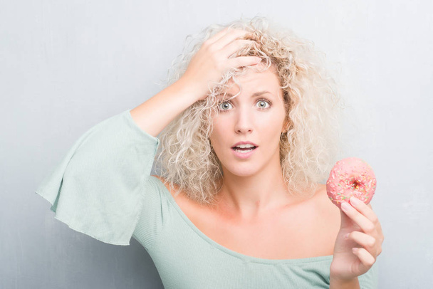 Young blonde woman over grunge grey background eating pink donut stressed with hand on head, shocked with shame and surprise face, angry and frustrated. Fear and upset for mistake. - Foto, immagini