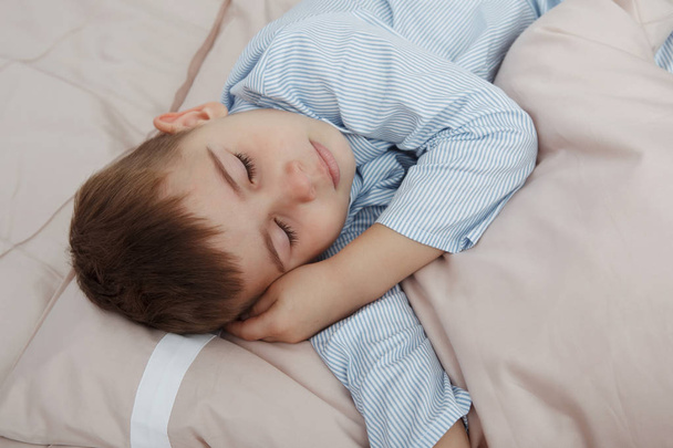 Kleiner Junge im blauen Schlafanzug liegt zusammengerollt in einem Ball und umarmt ein Kissen in einem Bett mit Bettwäsche. - Foto, Bild