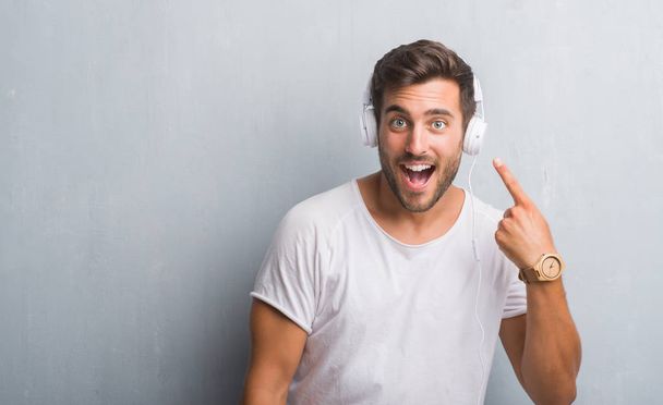Όμορφος νεαρός άνδρας πάνω από τοίχο γκρι grunge φορώντας ακουστικά να ακούτε μουσική πολύ χαρούμενος, δείχνοντας με το χέρι και δάχτυλα - Φωτογραφία, εικόνα