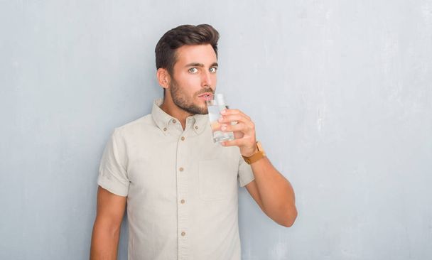 Knappe jongeman over grijs grunge wall drinkglas van water met een vertrouwen uitdrukking op slimme gezicht ernstige denken - Foto, afbeelding