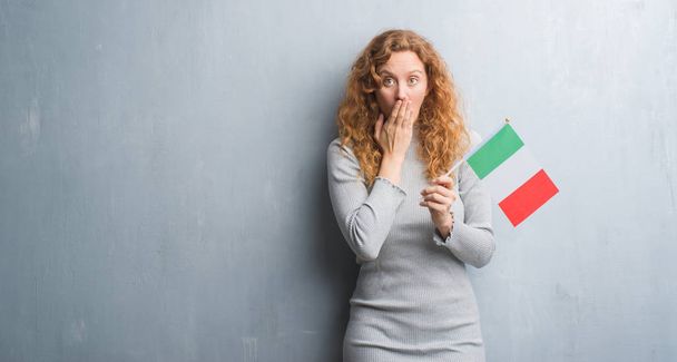 Ruda młoda kobieta ponad szary tło ściany trzymając flaga Włoch pokrywa usta ręką w szoku ze wstydem za błąd, wyrażenie strachu, przestraszony w milczeniu, tajne koncepcja - Zdjęcie, obraz