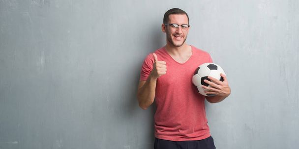 Kaukázusi fiatalember gazdaság labdarúgás labdarúgó szürke grunge falon boldog labda nagy mosollyal az ok jele ennek, hüvelykujját, ujjaival, kiváló jel - Fotó, kép