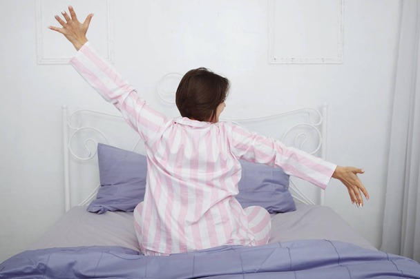 Nuori söpö tumma tukka nainen vaaleanpunainen pyjama istuu sängyllä tyynyt ja sininen vuodevaatteet. Taustakuva
. - Valokuva, kuva
