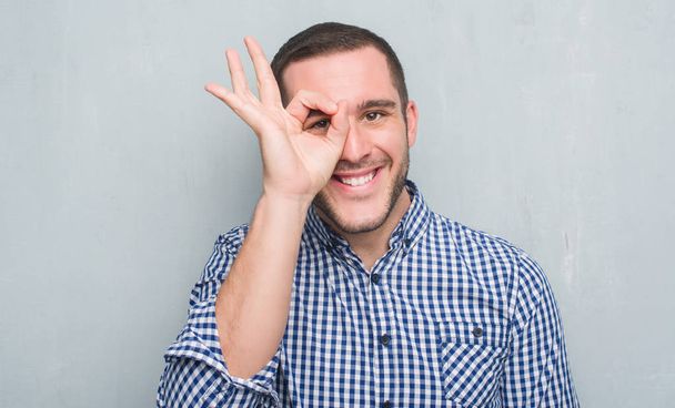Νεαρός Καυκάσιος πάνω από το τοίχο γκρι grunge με χαρούμενο πρόσωπο χαμογελά κάνει εντάξει σημάδι με το χέρι στο μάτι που αναζητούν μέσα από τα δάχτυλα - Φωτογραφία, εικόνα