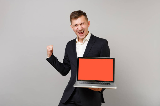 Радісний молодий бізнесмен у костюмі робить жест переможця, тримаючи комп'ютер ноутбука з порожнім порожнім екраном ізольовано на сірому фоні. Досягнення кар'єри багатства бізнес-концепції. Збільшити простір для копіювання
 - Фото, зображення