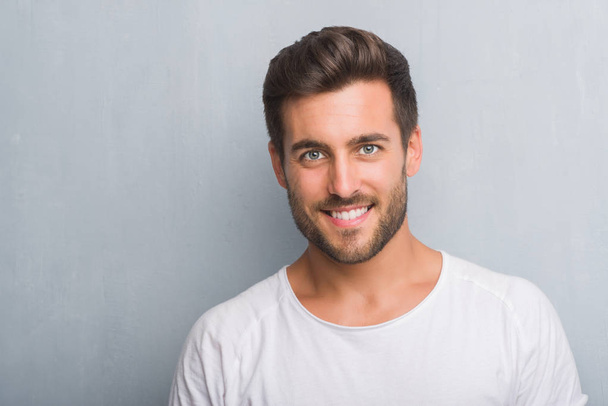 Schöner junger Mann über der grauen Grunge-Wand mit einem glücklichen Gesicht stehend und lächelnd mit einem selbstbewussten Lächeln, das Zähne zeigt - Foto, Bild
