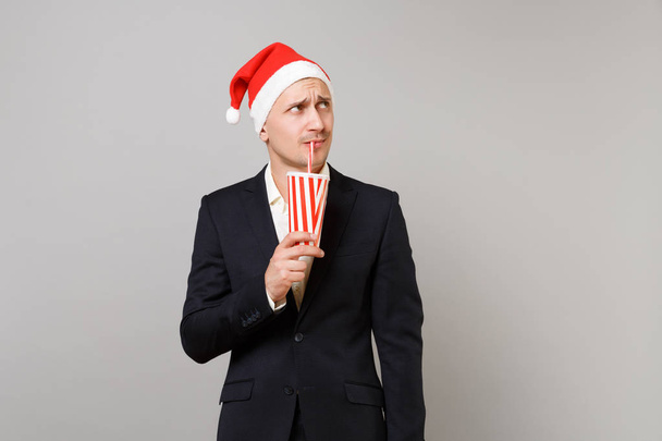 Puzzled business man in Christmas hat looking up, drinking soda from plastic cup isolated on grey background. Achievement career wealth business. Happy New Year 2019 celebration holiday party concept - Фото, изображение
