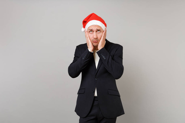 見上げるクリスマス帽子で疲れて動揺若いビジネスマンは、灰色の背景で隔離の頬に手を置きます。達成のキャリア豊富なビジネス。幸せな新しい年 2019年お祝い休日党概念 - 写真・画像
