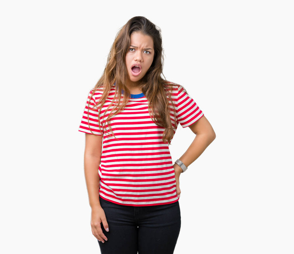 junge schöne brünette Frau trägt gestreiftes T-Shirt über isoliertem Hintergrund im Schockgesicht, sieht skeptisch und sarkastisch aus, überrascht mit offenem Mund - Foto, Bild