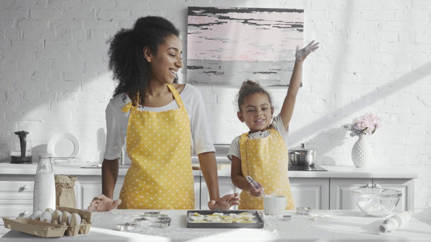 adorable african american child in apron having fun with flour while her mother standing near in kitchen - Footage, Video