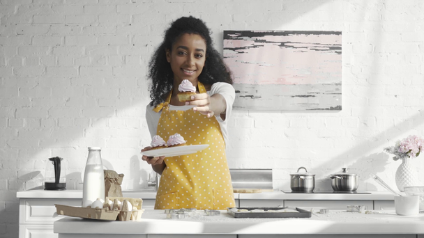 sonriente afroamericana joven en delantal sosteniendo plato y mostrando cupcakes en cocina
 - Imágenes, Vídeo