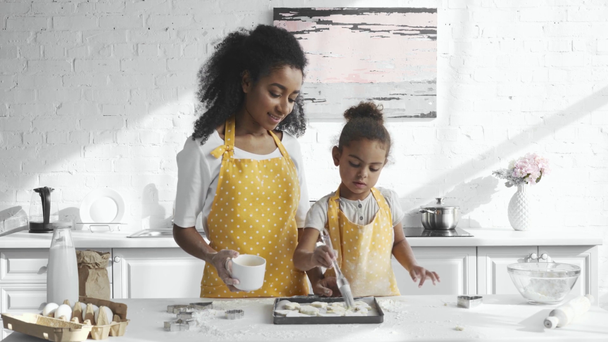 Konzentrierte afrikanisch-amerikanische Kind in Schürze Bürsten Teig auf Tablett, während ihre Mutter in der Nähe in der Küche stehen - Filmmaterial, Video