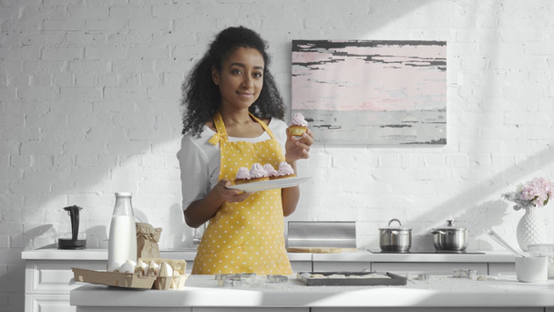 mulher americana africana alegre no avental segurando placa e mostrando cupcakes na cozinha
 - Filmagem, Vídeo