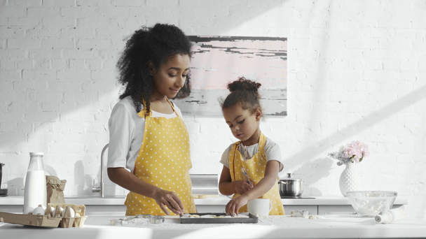 african american kobieta nauczanie jej córeczkę w fartuch szczotkowanie ciasta tacy w kuchni  - Materiał filmowy, wideo