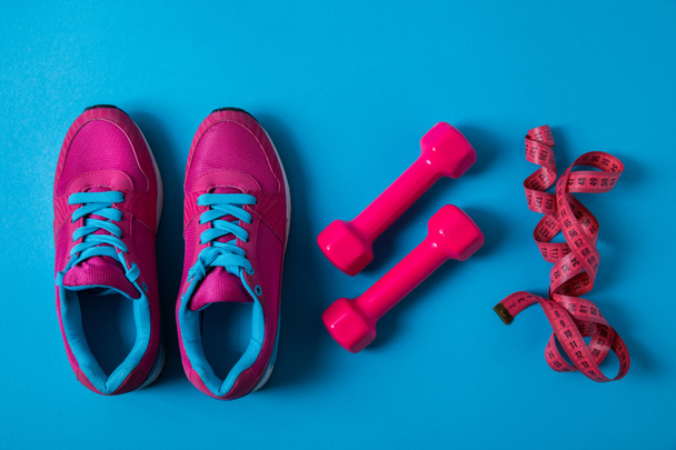 επίπεδη θέσει με τοποθετημένα ροζ αθλητικά παπούτσια, αλτήρες και μετροταινία τοποθετούνται σε σειρά που απομονώνονται σε μπλε, μινιμαλιστική έννοια  - Φωτογραφία, εικόνα