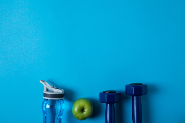elevated view of arranged sport bottle, green apple and dumbells isolated on blue, minimalistic concept  - Photo, Image