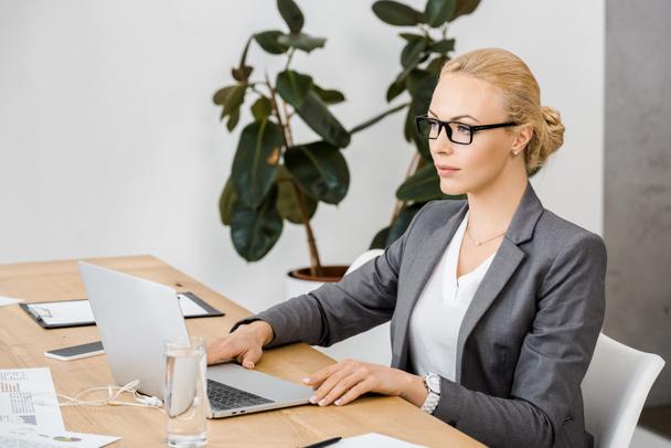jonge vrouw in glazen aan tafel zit en met behulp van laptop in kantoor - Foto, afbeelding