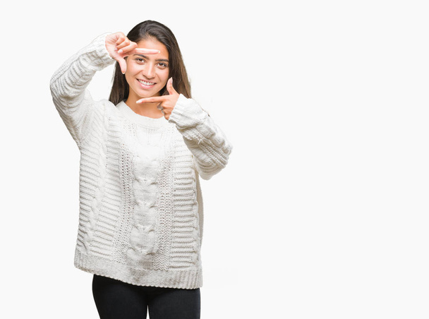 笑顔の幸せそうな顔で指と手作りフレーム分離の背景に冬のセーターを着た若い美しいアラブ女性。創造性と写真のコンセプト. - 写真・画像