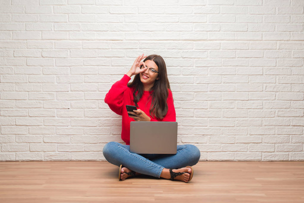 Young brunette woman sitting on the floor using laptop and smartphone with happy face smiling doing ok sign with hand on eye looking through fingers - Foto, afbeelding